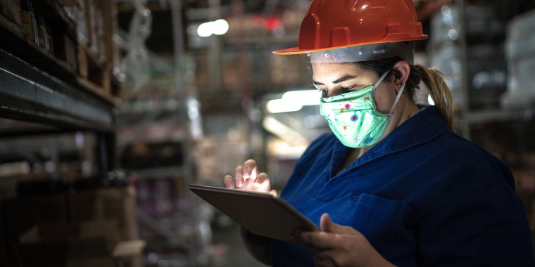 A woman in a mask and a hard hat works on a tablet.